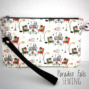 May include: A white zippered pouch with a black strap and a black zipper. The pouch has a repeating pattern of black cats reading books in green and pink chairs. The text "Paradise Falls SEWING" is printed on the bottom of the image.