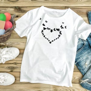 Puede incluir: Camiseta blanca con un dise&ntilde;o de mariposa negra en forma de coraz&oacute;n.