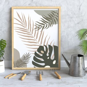 May include: A framed print featuring a white background with a stylised illustration of tropical leaves in shades of brown, green, and grey. The print is hanging on a grey wall with a watering can and potted plants in the foreground.