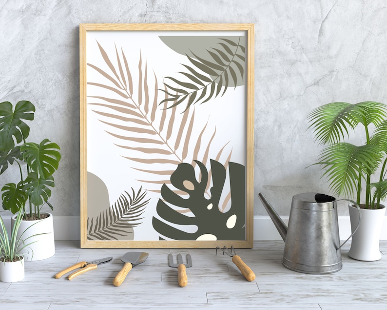 May include: A framed print featuring a white background with a stylised illustration of tropical leaves in shades of brown, green, and grey. The print is hanging on a grey wall with a watering can and potted plants in the foreground.