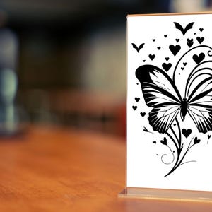 Puede incluir: Una silueta en blanco y negro de una mariposa con alas intrincadas y un dise&ntilde;o floral. La mariposa est&aacute; rodeada de peque&ntilde;os corazones.
