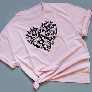 Puede incluir: Una camiseta rosa con un dise&ntilde;o de mariposa negra en forma de coraz&oacute;n.