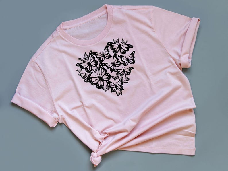 Puede incluir: Una camiseta rosa con un dise&ntilde;o de mariposa negra en forma de coraz&oacute;n.