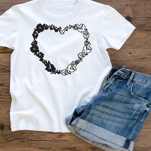 Puede incluir: Camiseta blanca con un dise&ntilde;o negro en forma de coraz&oacute;n hecho de mariposas. El coraz&oacute;n est&aacute; centrado en la camiseta. La camiseta est&aacute; sobre una superficie de madera con un par de pantalones cortos de mezclilla azul.