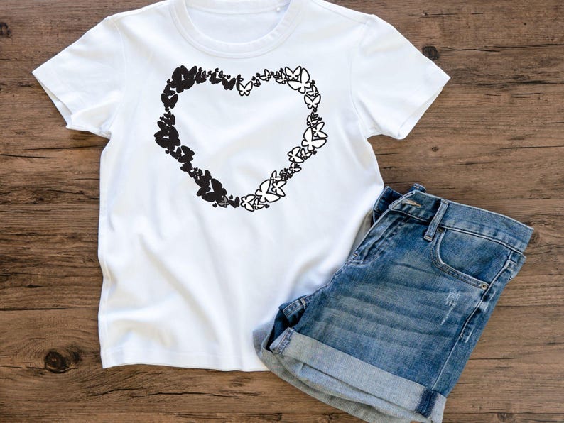 Puede incluir: Camiseta blanca con un dise&ntilde;o negro en forma de coraz&oacute;n hecho de mariposas. El coraz&oacute;n est&aacute; centrado en la camiseta. La camiseta est&aacute; sobre una superficie de madera con un par de pantalones cortos de mezclilla azul.