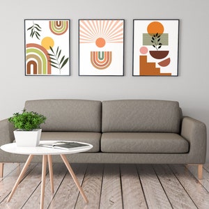 May include: Three framed abstract art prints featuring geometric shapes and colours in shades of brown, orange, yellow, green, and pink. The prints are hung on a white wall above a grey sofa.
