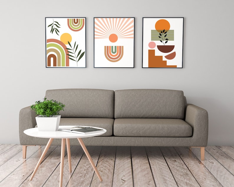 May include: Three framed abstract art prints featuring geometric shapes and colours in shades of brown, orange, yellow, green, and pink. The prints are hung on a white wall above a grey sofa.