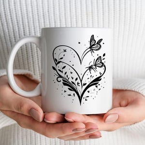 Puede incluir: Taza de caf&eacute; de cer&aacute;mica blanca con un dise&ntilde;o de silueta negra de un coraz&oacute;n con dos mariposas y detalles florales.