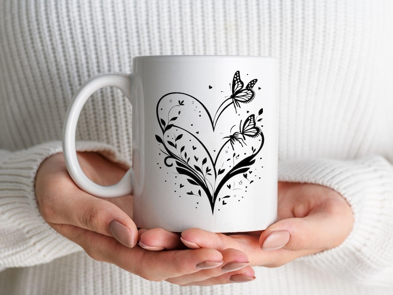 Puede incluir: Taza de caf&eacute; de cer&aacute;mica blanca con un dise&ntilde;o de silueta negra de un coraz&oacute;n con dos mariposas y detalles florales.
