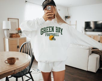 May include: White hoodie with the text "GREEN BAY FOOTBALL" in green lettering and a yellow football helmet graphic. The person in the image is wearing a black baseball cap.