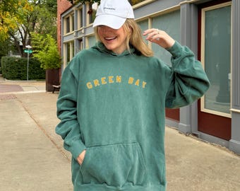 May include: A teal green hoodie with the text "GREEN BAY" in yellow block letters. The hoodie has a front pocket and is paired with light blue jeans and a white baseball cap.