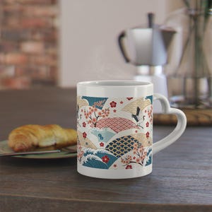 Könnte beinhalten: Weiße Keramiktasse mit herzförmigem Griff, mit einem japanisch inspirierten Design. Das Design umfasst Wellen, Kirschblüten, Kraniche und geometrische Muster in Rot, Blau und Gold. Ein Croissant liegt auf einem Teller in der Nähe.