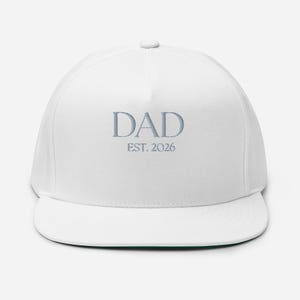 May include: White flat-brimmed baseball cap with the word "DAD" embroidered in light blue, along with "EST. 2026". The cap has a structured crown and a flat brim, suitable for casual wear.