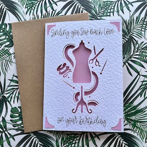 May include: A birthday card with a pink dress form design and the words "Sending you Sew much love on your birthday." The card is white with a textured surface and comes with a brown envelope.