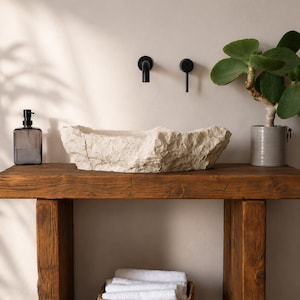 Peut inclure: Un meuble-lavabo rustique avec un évier en pierre naturelle. L'évier est de couleur beige clair et présente une surface rugueuse et texturée. Le meuble est en bois brun foncé et possède une étagère inférieure avec un panier tressé contenant des serviettes blanches. Une plante en pot et des accessoires noirs complètent la scène.