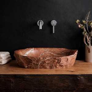 May include: A red stone sink with white veining, featuring a rough, natural texture, is displayed on a wooden surface. Chrome faucets are mounted above. A stack of white towels and a potted plant are in the background.