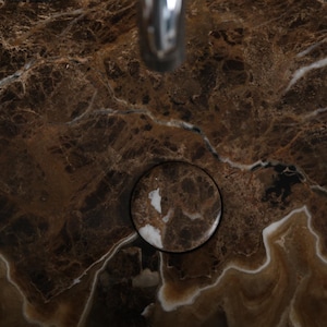May include: Close-up of a brown marble surface with white veining. A circular drain is visible, reflecting the marble pattern. The image showcases the natural patterns and textures of the stone, suitable for bathroom or kitchen design.