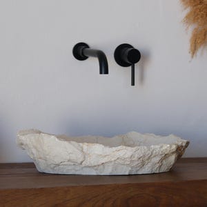 May include: A textured, off-white stone-like sink sits on a wooden surface, complemented by a black faucet and a white pitcher with dried plants. A woven basket with white towels is on the left.