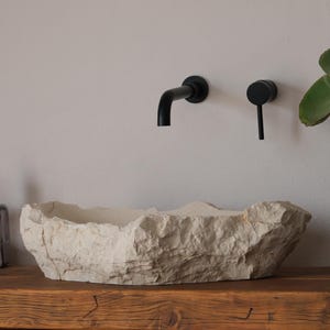 May include: A rustic bathroom sink made of light-colored stone sits on a wooden shelf. Black faucet fixtures are mounted on the wall above the sink. A potted plant with large green leaves adds a natural touch to the space.