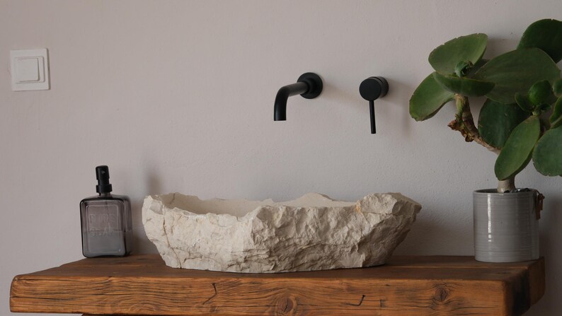 May include: A rustic bathroom sink made of light-colored stone sits on a wooden shelf. Black faucet fixtures are mounted on the wall above the sink. A potted plant with large green leaves adds a natural touch to the space.