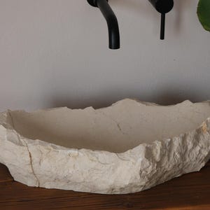 May include: A natural stone sink with a rough, textured exterior. The off-white basin has an irregular shape and sits on a wooden surface. Black faucet fixtures are mounted above the sink. A potted plant is visible in the background.