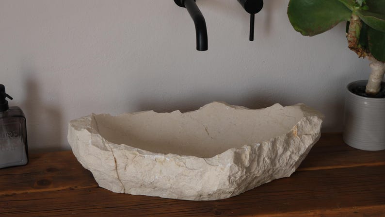 May include: A natural stone sink with a rough, textured exterior. The off-white basin has an irregular shape and sits on a wooden surface. Black faucet fixtures are mounted above the sink. A potted plant is visible in the background.