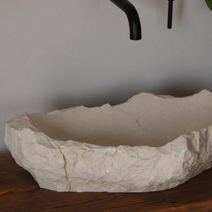 May include: A natural stone vessel sink with a rough, textured exterior and a smooth interior. The sink is off-white and sits on a wooden surface. A black faucet and soap dispenser are visible.