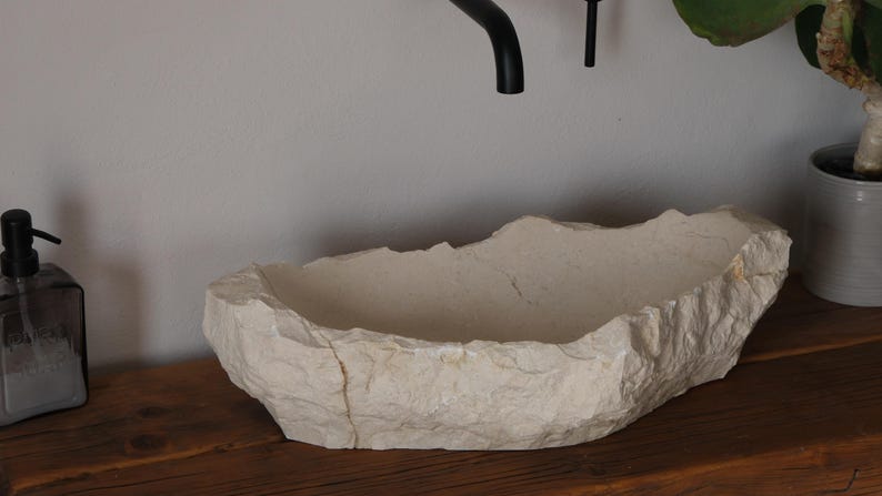 May include: A natural stone vessel sink with a rough, textured exterior and a smooth interior. The sink is off-white and sits on a wooden surface. A black faucet and soap dispenser are visible.