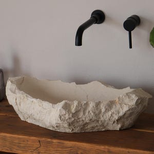 May include: A textured, off-white stone basin sits on a rustic wooden table. Black faucet fixtures are mounted on the wall above the basin. A clear bottle with a black pump and a potted plant are also on the table.