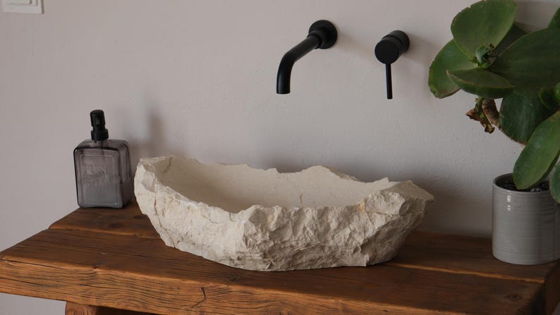 May include: A textured, off-white stone basin sits on a rustic wooden table. Black faucet fixtures are mounted on the wall above the basin. A clear bottle with a black pump and a potted plant are also on the table.