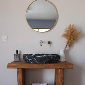 May include: A black marble vessel sink with white veining sits on a rustic wooden vanity table. Above the sink, a round gold-framed mirror is mounted on the wall. A white pitcher with dried grass, a soap dispenser, and white towels are present.