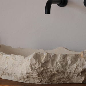 May include: A natural stone-like bathroom sink with a rough, textured exterior. The off-white basin sits on a wooden surface, with black fixtures and a soap dispenser visible. The sink's design evokes a rustic, organic aesthetic.