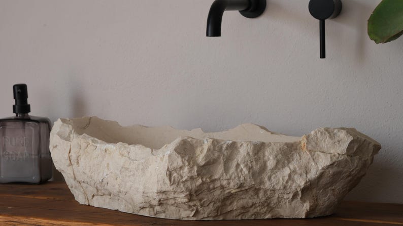 May include: A natural stone-like bathroom sink with a rough, textured exterior. The off-white basin sits on a wooden surface, with black fixtures and a soap dispenser visible. The sink's design evokes a rustic, organic aesthetic.