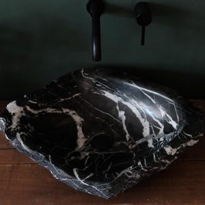 Puede incluir: Un lavabo de mármol blanco y negro con un borde áspero y natural. El lavabo está colocado sobre una encimera de madera.