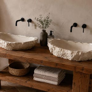May include: Two stone-like sinks with rough edges sit atop a rustic wooden vanity. Black faucets and a small vase with greenery are mounted on the wall. A woven basket and folded towels are on the vanity shelf. The sinks are a light beige color.
