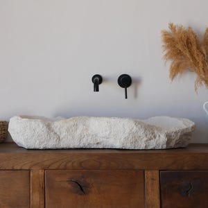 Puede incluir: Un lavabo de baño de piedra natural con una superficie rugosa y texturizada se encuentra sobre un mueble de madera oscura. Los grifos negros están montados en la pared sobre el lavabo. Una jarra blanca con plantas secas y una cesta de mimbre con toallas blancas completan la escena.