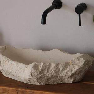 May include: A beige, stone-like bathroom sink with a rough, textured surface sits on a wooden countertop. Black faucet fixtures are mounted on the wall above the sink. A soap dispenser and a potted plant are also visible.