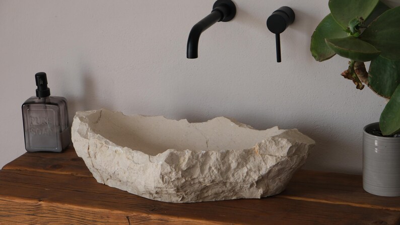 May include: A beige, stone-like bathroom sink with a rough, textured surface sits on a wooden countertop. Black faucet fixtures are mounted on the wall above the sink. A soap dispenser and a potted plant are also visible.