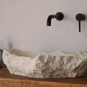 May include: A stone-like, off-white bathroom sink with a rough, textured surface sits on a wooden countertop. Black faucet and handle fixtures are mounted on the wall above. A glass bottle and a plant are also visible.