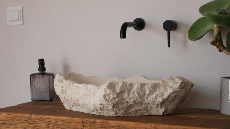 May include: A stone-like, off-white bathroom sink with a rough, textured surface sits on a wooden countertop. Black faucet and handle fixtures are mounted on the wall above. A glass bottle and a plant are also visible.