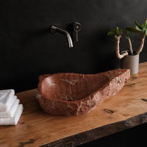 May include: A natural, rough-hewn red marble vessel sink with a polished interior basin. The sink features irregular, jagged exterior edges and white veining throughout the stone. It is positioned on a wooden surface next to white folded towels.