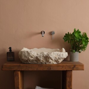 May include: A rustic bathroom vanity with a stone sink. The sink is a light beige color and sits on a wooden table. A green plant in a gray pot and a dark soap dispenser are on the table. White towels are in a woven basket.
