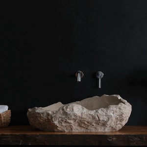 May include: A natural stone vessel sink with a rough, textured exterior sits on a wooden surface. The sink is beige and has an organic shape. A woven basket with white towels and a potted plant are on either side.