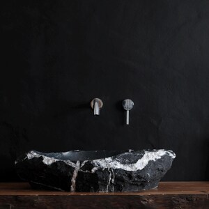 May include: A black and white stone vessel sink with a natural, organic shape. The sink is mounted on a wooden surface against a black wall. Two silver faucets are mounted on the wall above the sink. A white pitcher with dried plants is on the right.
