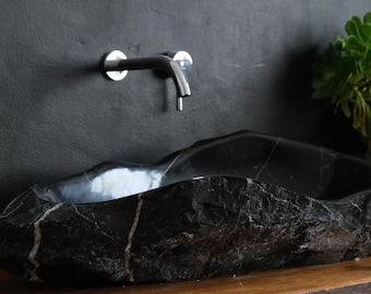 May include: A black stone sink with a natural, rough exterior and a smooth interior. The sink is mounted below a chrome faucet against a dark gray wall. A green plant in a white vase sits on a wooden surface.