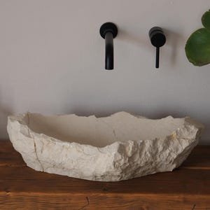 May include: A natural stone-like bathroom sink with a rough, textured exterior. The sink is off-white and sits on a wooden surface. Black faucets are mounted on the wall above. A potted plant and soap dispenser are also present.