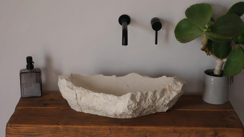 May include: A natural stone-like bathroom sink with a rough, textured exterior. The sink is off-white and sits on a wooden surface. Black faucets are mounted on the wall above. A potted plant and soap dispenser are also present.