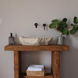 May include: A rustic bathroom vanity with a stone sink, black fixtures, and a wooden base. A gray glass soap dispenser and a potted plant with green leaves add to the decor. A woven basket with white towels sits on the lower shelf.