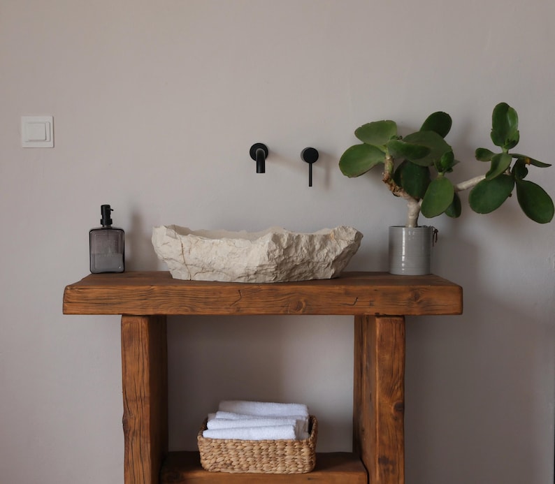 May include: A rustic bathroom vanity with a stone sink, black fixtures, and a wooden base. A gray glass soap dispenser and a potted plant with green leaves add to the decor. A woven basket with white towels sits on the lower shelf.