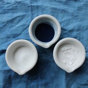 Peut inclure: Trois bols en céramique blanche sur un fond de tissu bleu. Un bol contient de la poudre bleue, un autre de la poudre blanche et le troisième est vide.